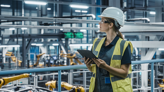 Female Car Factory Engineer in High Visibility Vest Using Tablet Computer. Automotive Industrial Manufacturing Facility Working on Vehicle Production with Robotic Technology. Automated Assembly Plant.