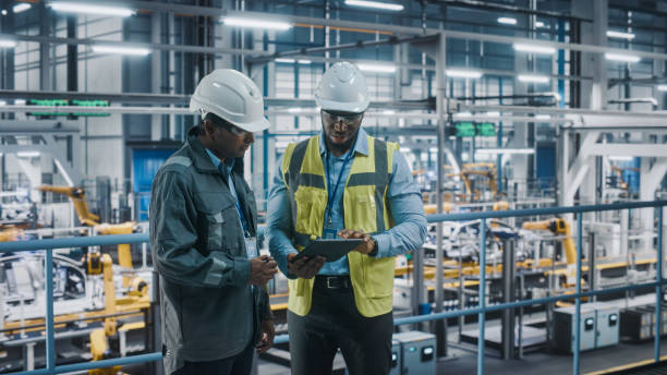 istock Multiethnic Manager and African American Car Factory Engineer in Uniform Using Tablet Computer. Automotive Industry 4.0 Manufacture Employees Discuss Work on Vehicle Assembly Plant. 1352428854