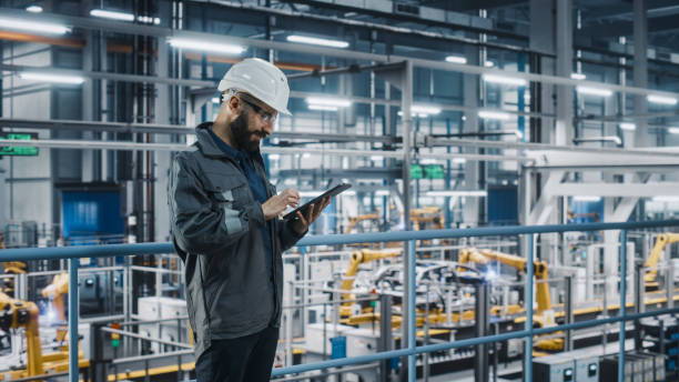 istock Car Factory Engineer in Work Uniform Using Tablet Computer. Automotive Industry 4.0 Manufacturing Facility Working on Vehicle Production with Robotic Arms Technology. Automated Assembly Plant. 1352428886