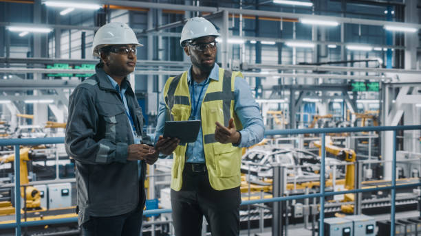 istock Multiethnic Manager and African American Car Factory Engineer in Uniform Using Tablet Computer. Automotive Industry 4.0 Manufacture Employees Discuss Work on Vehicle Assembly Plant. 1352428892