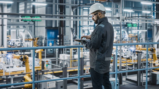 istock Car Factory Engineer in Work Uniform Using Laptop Computer. Automotive Industrial Manufacturing Facility Working on Vehicle Production with Robotic Arms Technology. Automated Assembly Plant. 1352428898