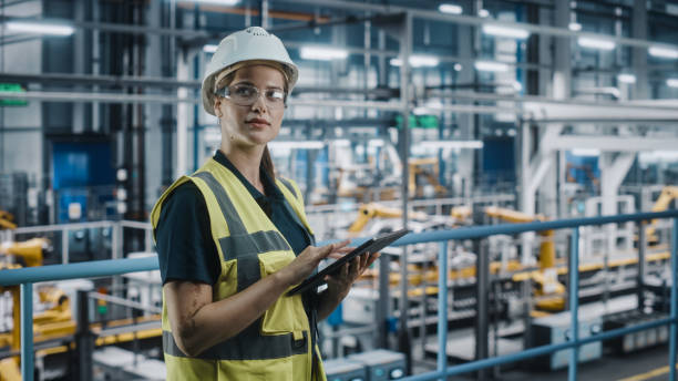 istock Female Car Factory Engineer in High Visibility Vest Using Laptop Computer. Automotive Industrial Manufacturing Facility Working on Vehicle Production with Robotic Arms. Automated Assembly Plant. 1352428921