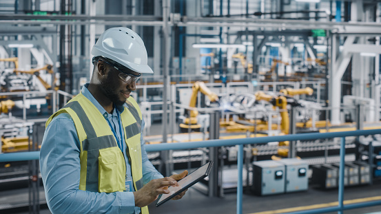 Feliz ingeniero afroamericano de fábrica de automóviles en chaleco de alta visibilidad usando tableta. Instalación industrial automotriz que trabaja en la producción de vehículos en la planta de ensamblaje de tecnología automatizada. photo