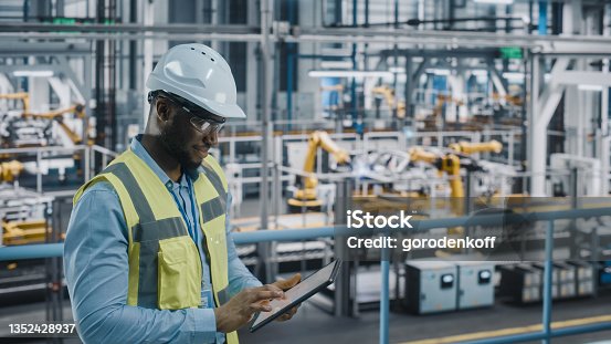 istock Happy African American Car Factory Engineer in High Visibility Vest Using Tablet Computer. Automotive Industrial Facility Working on Vehicle Production on Automated Technology Assembly Plant. 1352428937