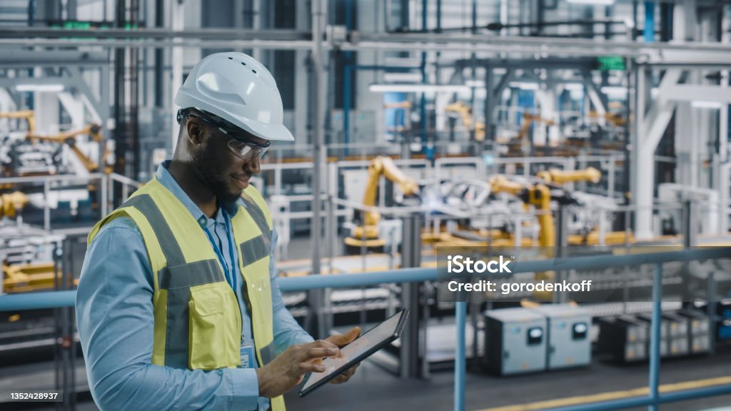 Happy African American Car Factory Engineer in High Visibility Vest Using Tablet Computer. Automotive Industrial Facility Working on Vehicle Production on Automated Technology Assembly Plant. Manufacturing Stock Photo