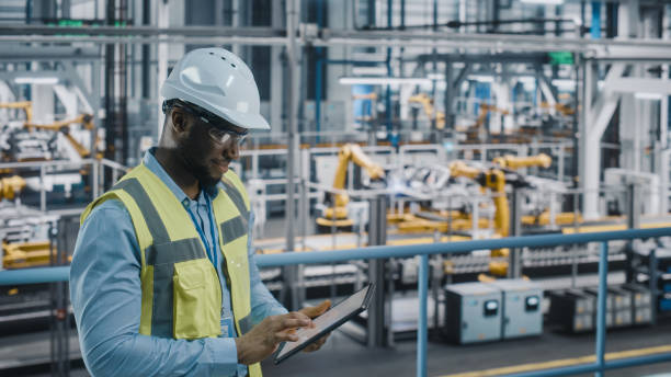 happy african american car factory engineer in high visibility vest using tablet computer. automotive industrial facility working on vehicle production on automated technology assembly plant. - löpande band bildbanksfoton och bilder