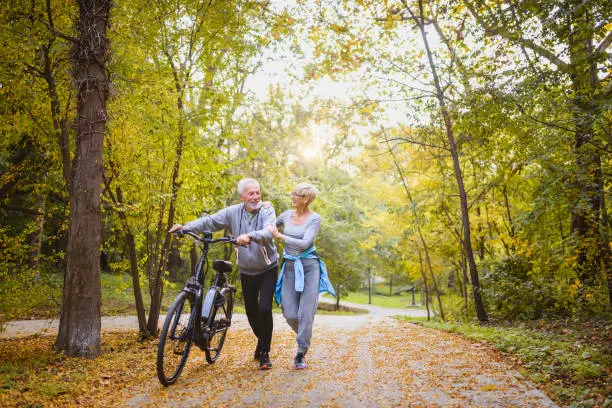 Photo of Cheerful active senior couple with bicycles walking through park together. Perfect activities for elderly people.