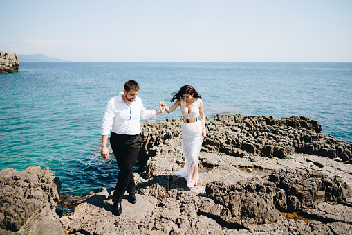 The bride and groom walk holding hands on the rocky shore . High quality photo