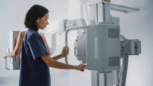 Photo of Hospital Radiology Room: Beautiful Multiethnic Woman Standing Topless Next to X-Ray Machine while Female Latin Nurse Adjusts it. Healthy Patient Undergoes Medical Exam Scanning with the Doctor's Help.