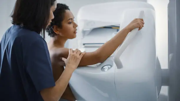 Friendly Female Doctor Explains the Mammogram Procedure to a Topless Latin Female Patient with Curly Hair Undergoing Mammography Scan. Healthy Female Does Cancer Prevention Routine in Hospital Room.