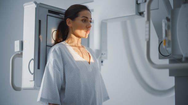 Hospital Radiology Room: Beautiful Multiethnic Woman Standing in Medical Gown in the X-Ray Machine. Adult Female Undergoes Healthcare Exam and is Scanning Chest, Heart, Lungs in Modern Clinic Office. sala de radiología del hospital: hermosa mujer multiétnica de pie con bata médica en la máquina de rayos x. la mujer adulta se somete a un examen de atención médica y está escaneando el tórax, el corazón y los pulmones en la oficina de la clínica