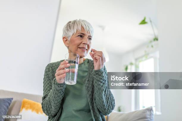 Smiling Mature Woman Relieving Disease Symptoms Taking A Painkiller Pill Sitting On Sofa Of In Living Room Stock Photo - Download Image Now
