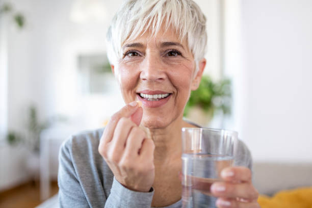 smiling happy healthy middle aged 50s woman holding glass of water taking dietary supplement vitamin pill. old women multivitamins antioxidants for anti age beauty. - bottle vitamin pill nutritional supplement white imagens e fotografias de stock