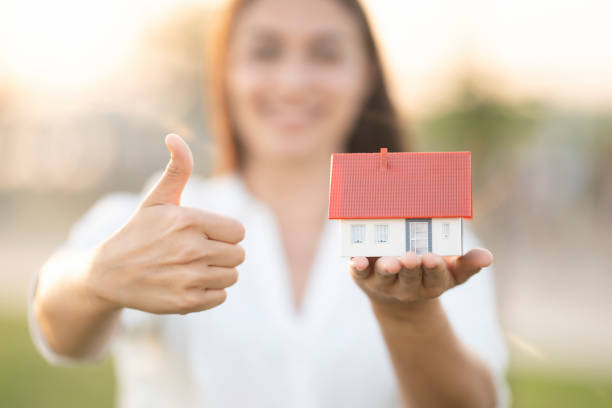 Hand is holding a model house. Another one thumbs up. She is smiling.