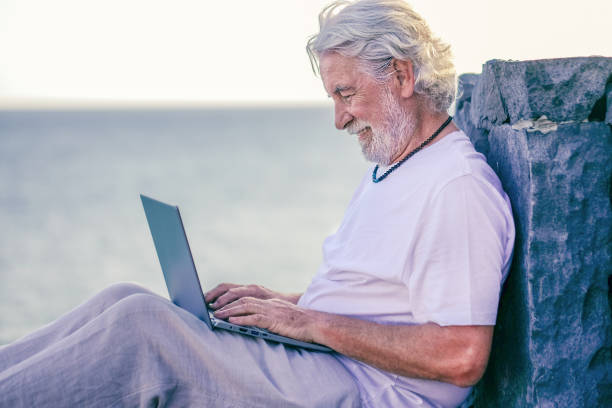 istock Smiling senior white-haired man sitting by the sea using laptop computer. Relaxed elderly man wearing necklace enjoying tech and social 1352444216