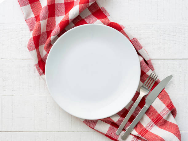 istock Directly overhead view of an empty white ceramic dinner plate on a red and white checkered gingham table cloth on a rustic white wood table background with a knife and fork next to the plate. 1352445037