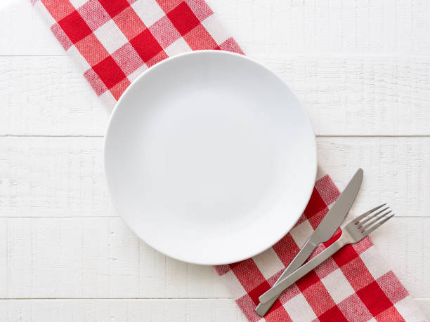 Directly overhead view of an empty white ceramic dinner plate on a red and white checkered gingham table cloth on a rustic white wood table background with a knife and fork next to the plate.