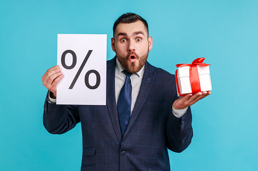 Wow, huge discounts! Astonished businessman wearing dark official style suit showing to camera percent mark and gift box, surprised with bonuses. Indoor studio shot isolated on blue background.