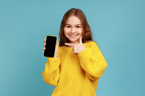 Portrait of little girl pointing finger at blank screen of mobile phone, looking smiling to camera, wearing yellow casual style sweater. Indoor studio shot isolated on blue background.