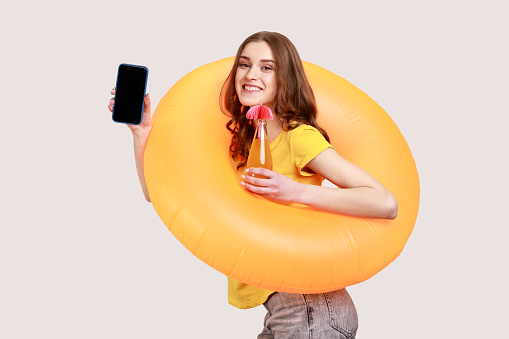 Portrait of smiling curly haired teenage girl in yellow casual style T-shirt with rubber ring, ready for vacation, showing blank screen of smart phone. Indoor studio shot isolated on gray background.