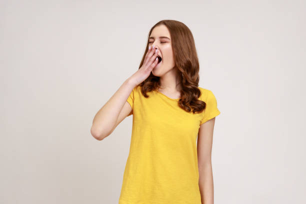porträt eines schläfrigen teenager-mädchens mit welligem haar in lässigem gelbem t-shirt, das mit geschlossenen augen gähnt und den mund bedeckt und sich erschöpft fühlt, ruhe braucht. - yawning stock-fotos und bilder