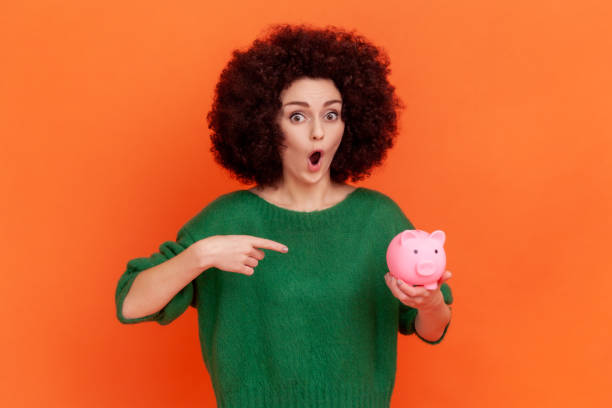 Portrait of shocked woman with Afro hairstyle wearing green casual style sweater standing pointing at piggy bank in her hand, advantageous bank offer. Portrait of shocked woman with Afro hairstyle wearing green casual style sweater standing pointing at piggy bank in her hand, advantageous bank offer. Indoor studio shot isolated on orange background. a penny saved stock pictures, royalty-free photos & images