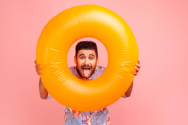 portrait of brunette bearded crazy excited man holding orange rubber ring in hands, looking at camera with funny expression, showing tongue out. - party beach indian ethnicity adult imagens e fotografias de stock