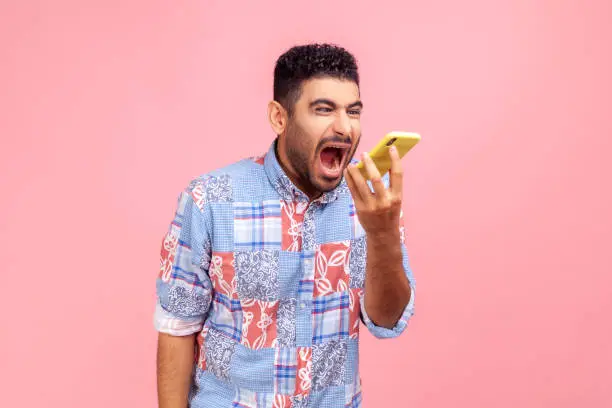 Portrait of angry mad man with beard in blue casual shirt loudly screaming talking phone, leaving voice message with furious aggressive expression. Indoor studio shot isolated on pink background.