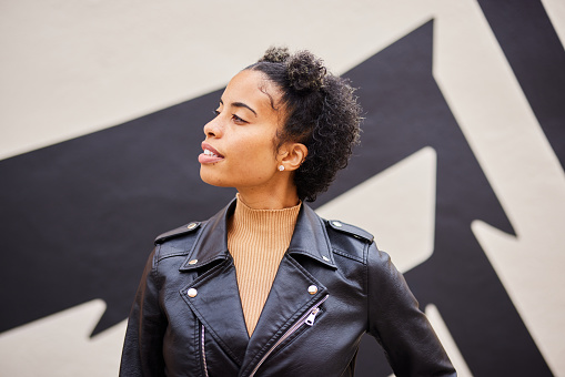 Young woman wearing a leather jacket looking off in the distance while standing outside in front of a wall mural