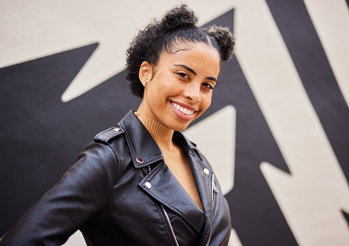 Portrait of a smiling young woman wearing a leather jacket standing outside in front of a wall mural