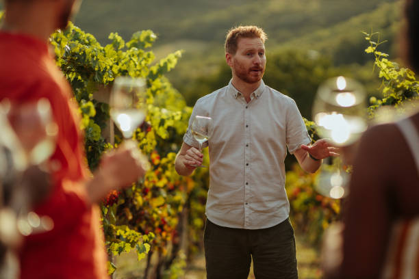 Happy wine tourists tasting wine in vineyard Group of adults during wine tasting in a vineyard. Ginger man presenting wine and holding glass of wine winery stock pictures, royalty-free photos & images