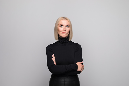 Portrait of beautiful mature woman wearing black turtleneck, standing with arms crossed and looking away. Studio shot, grey background.