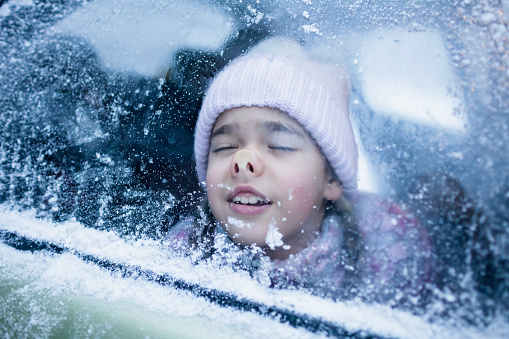 Little girl wearing in winter clothes pressed her face against car window glass funny flattening nose, family road trip in winter, active weekend