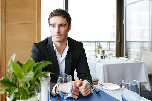 Serious Man in suit which sitting by the table in restaurant and looking away