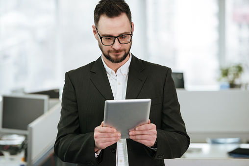 Pensive Bearded business man in suit and eyeglasses which using tablet computer in office