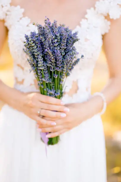 Bride in a white lace dress holds a lavender bouquet tied with a lilac ribbon. Close-up. High quality photo