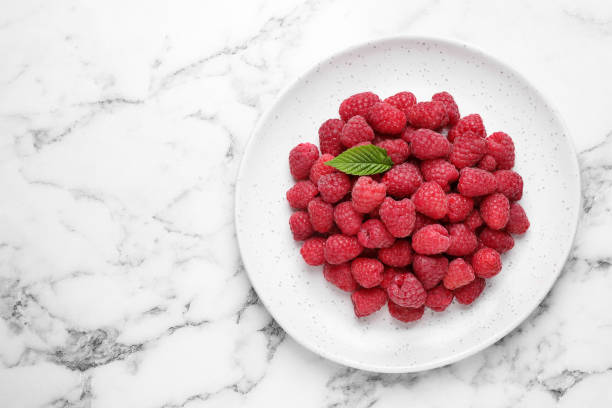 istock Delicious fresh ripe raspberries in plate on white marble table, top view. Space for text 1352473060