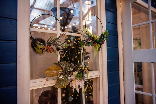 Round stylish wreaths hang on the window of the wooden blue house. Christmas tree with gifts is visible through the glass. New year home decoration. Trending decorations from natural objects.