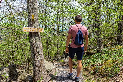 One young man with drawstring bag backpack walking on Wintergreen ski resort village town nature Highlands leisure hiking trail in forest of Virginia in spring with sign