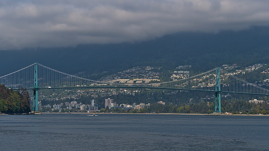Beautiful view of Lions Gate Bridge (First Narrows Bridge) crossing Burrard Inlet in Vancouver, British Columbia, Canada with the city of West Vancouver in background and clouds above the hills.