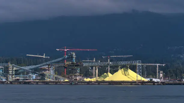 Photo of View of huge stock pile of bright yellow colored Sulphur (S) at bulk cargo terminal in North Vancouver, Canada with conveyor belts and cranes in front of cloud-covered hills.