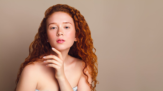 portrait of red-haired girl with long curly hair and cute freckles on beige background in the studio. Retouched photo. Skin care concept with pigmentation