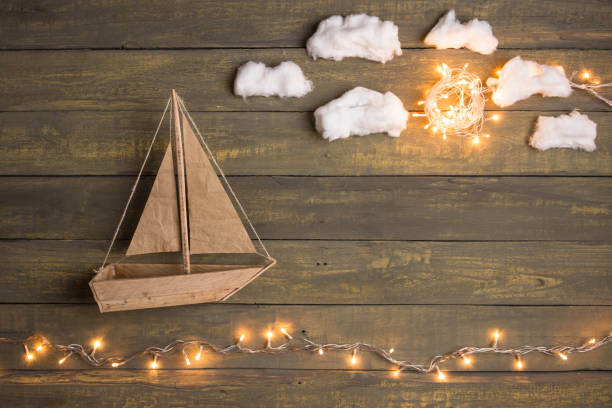 Travel and adventure creative concept - toy boat on a wooden background with cotton clouds. Christmas lights as a sea waves stock photo