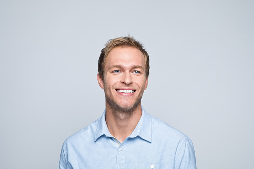 Portrait of handsome young man wearing blue shirt, looking away and smiling. Studio shot of male entrepreneur against grey background.
