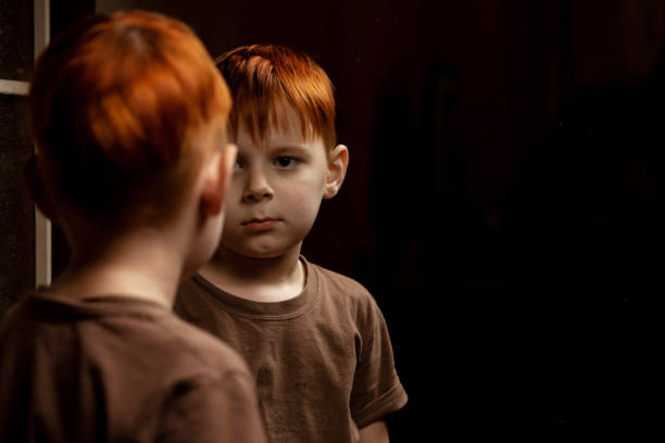 A young red-haired boy looks thoughtfully at himself in the mirror on a dark background. Childhood crisis, depression concept. Place for your text.