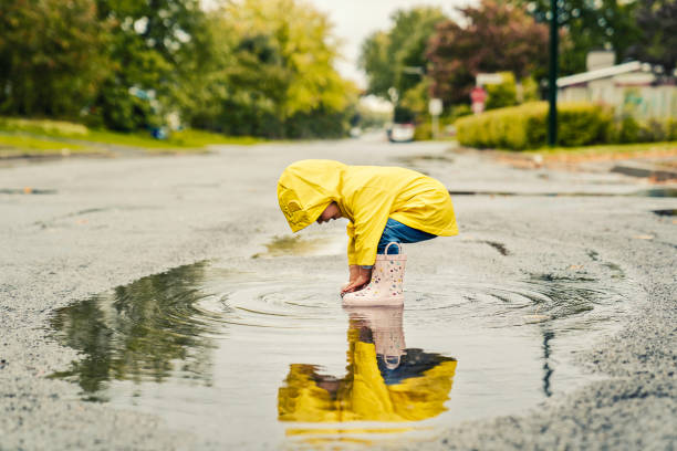 divertida y linda niña con abrigo amarillo impermeable y botas jugando bajo la lluvia - puddle fotografías e imágenes de stock