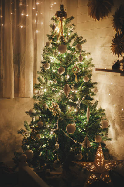 istock Stylish Christmas tree decorated with modern white baubles, boho ornaments and golden lights. Merry Christmas! Atmospheric festive scandinavian room. Christmas eve. Magic time. Space for text 1352498711