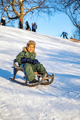 Little afro-american boy in warm overalls, hat and dumplings having fun riding sled from mountain hill slide at high speed on sunny winter day at sunset in park or forest. Active, leisure, holidays