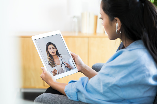 A serious young woman listens attentively as a female physician talks to her during a telemedicine appointment. The young woman is using a digital tablet to participate in the appointment.