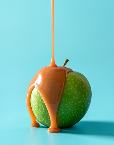 Pouring melted caramel sauce on a green apple, isolated on a blue background. Delicious caramel apple on a blue table.